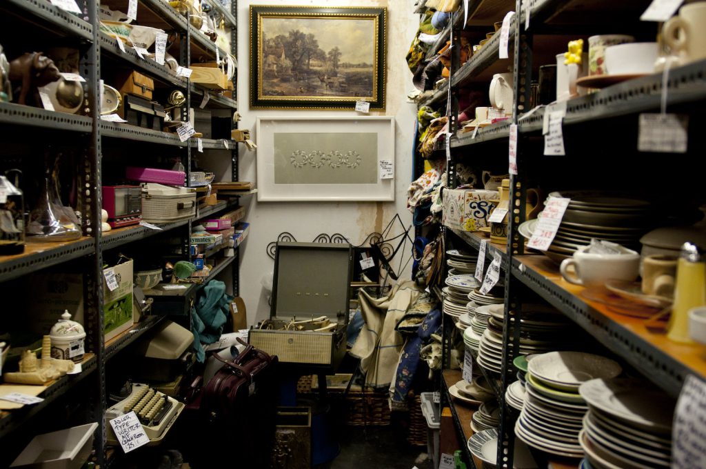 Two shelving units with multiple levels are packed with an assortment of objects. More objects are on the ground below two paintings hung on the back wall.
