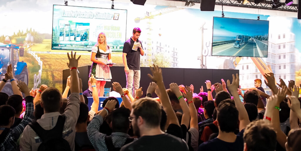 A group of people, some raising their hands, gather in front of a stage where a woman in traditional German dress and a man wearing white pants and a black shirt speak in front of video game displays.