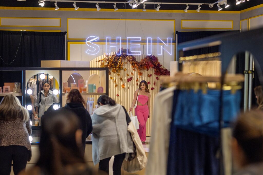 People wearing brown and tan coats shop in front of a woman in a pink outfit beneath a neon blue sign that reads SHEIN (pronounced “she-in”).