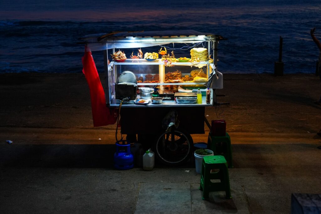 A transparent display case atop a cart glows against fading beachside evening light. Flatbreads and other local delicacies are in the case.