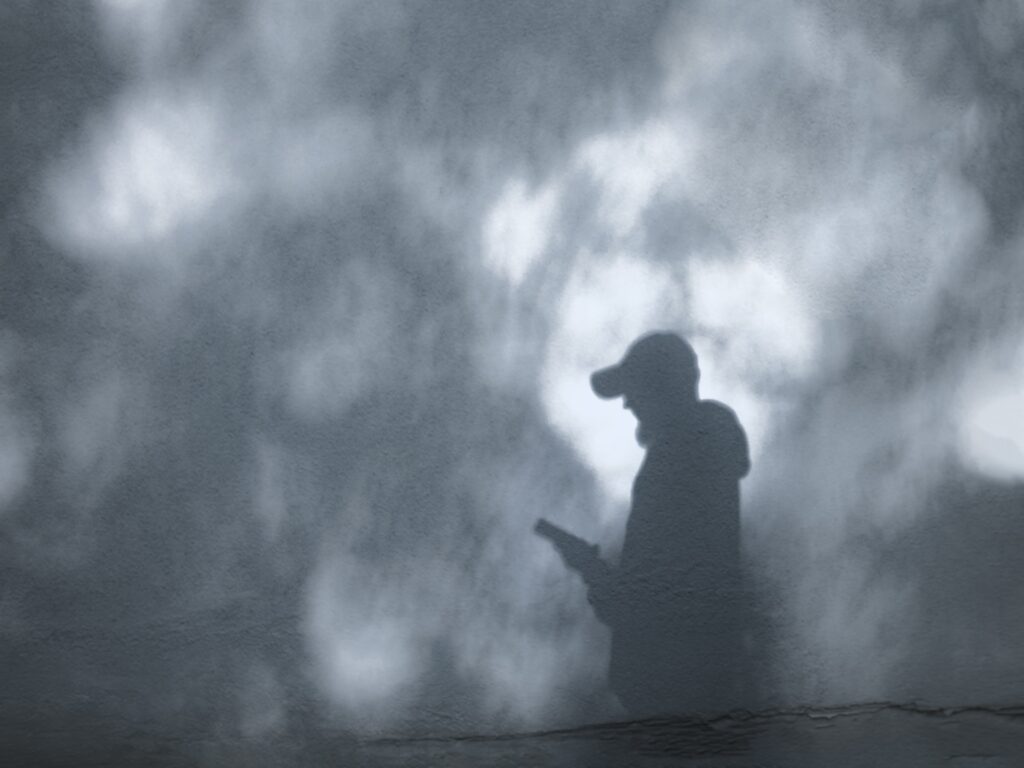 A shadow of a person in a baseball cap holding a smartphone is cast against a gray wall.