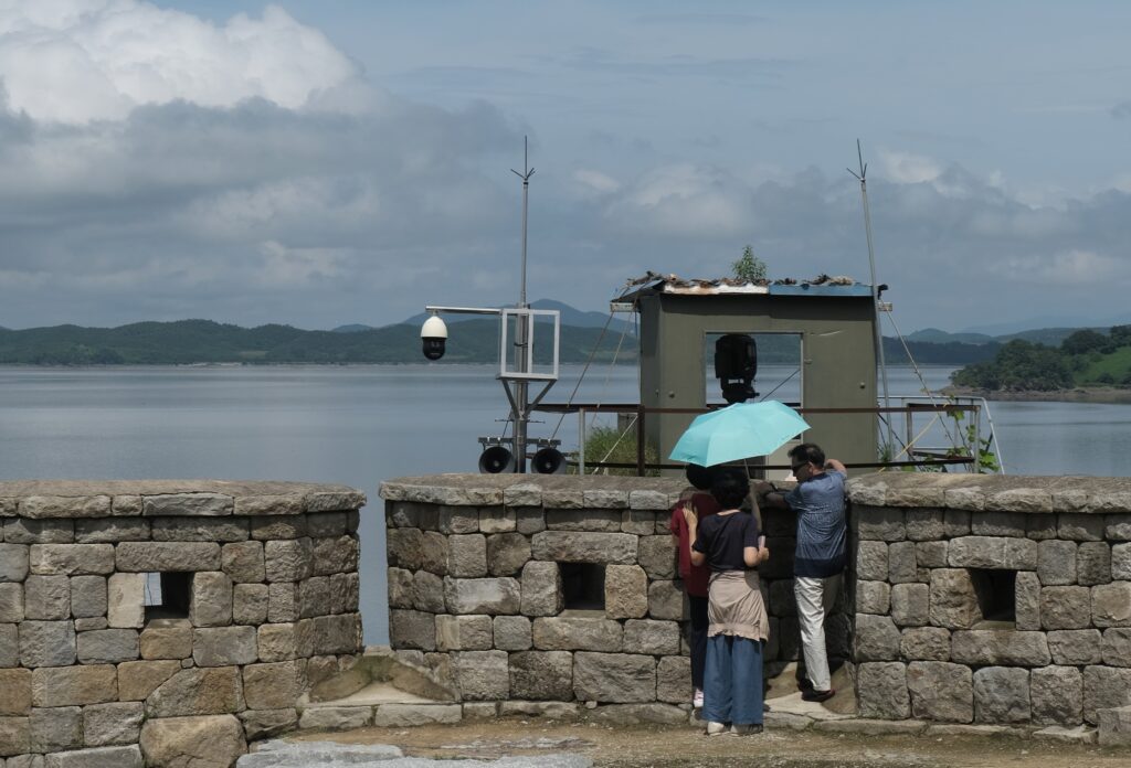 Standing at a lookout spot adjacent to a stone wall, a small group holding a blue umbrella looks across a large body of water at the opposite shore.